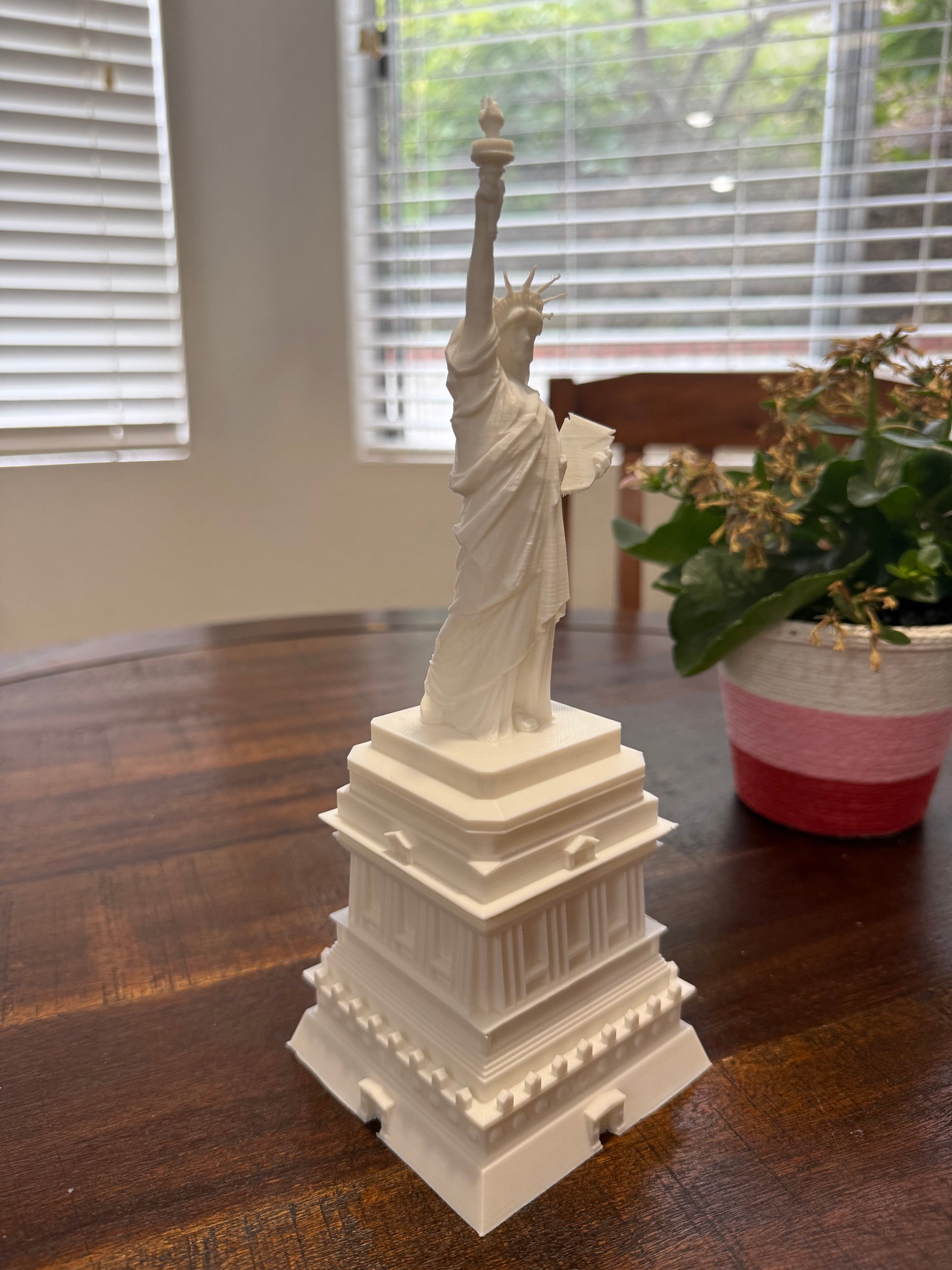 3D printed Statue of Liberty placed on a round wooden table with a plant in the background, showcasing detail and base.